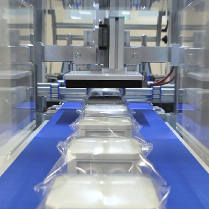 Zbliżenie na Automat pakujący poziomy FLOWPACK SBM550S zgrzewający kilka prostokątnych produktów w przezroczystych plastikowych opakowaniach na niebieskim przenośniku taśmowym w warunkach fabrycznych.