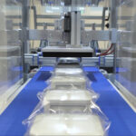 Zbliżenie na Automat pakujący poziomy FLOWPACK SBM750S zgrzewający prostokątne opakowania z przezroczystej folii plastikowej na niebieskim przenośniku, podkreślający jego wydajny zautomatyzowany proces pakowania.