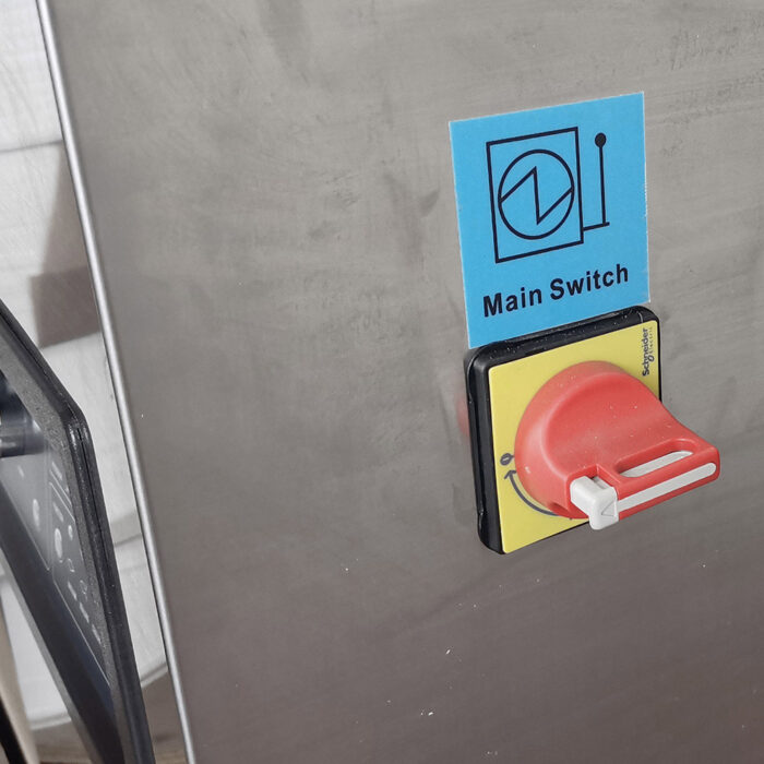Zbliżenie czerwono-żółtego wyłącznika głównego z białym uchwytem na metalowym panelu, oznaczonego jako "Wyłącznik główny" z symbolem elektrycznym powyżej, obok Komorowej pakowarki próżniowej Eco Vac 400D TB.