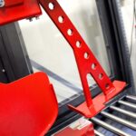 Zbliżenie jaskrawoczerwonego metalowego wspornika z otworami, zamontowanego na Automatycznej zaklejarce Auto Tape 50 HM w pobliżu okna lub obudowy bezpieczeństwa, z odblaskowymi metalowymi listwami w tle.