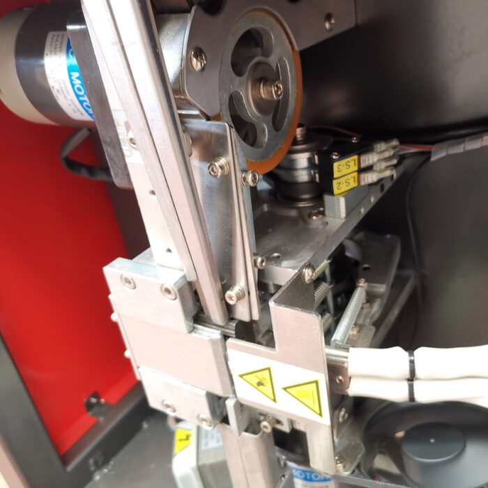 Zbliżenie Półautomatyczna bandownica do palet Pallet Strap T45, pokazująca metalowe koła zębate, przewody i etykiety bezpieczeństwa wewnątrz czerwono-srebrnej obudowy; komponenty maszyny wydają się być przeznaczone do przenoszenia lub zabezpieczania taśm paletowych.