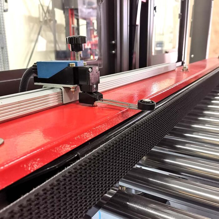Zbliżenie automatycznej zaklejarki Auto Tape 50 CF z czerwoną metalową ramą, niebiesko-czarnym pokrętłem regulacji i ramieniem pomiarowym nad przenośnikiem metalowych rolek w otoczeniu przemysłowym.