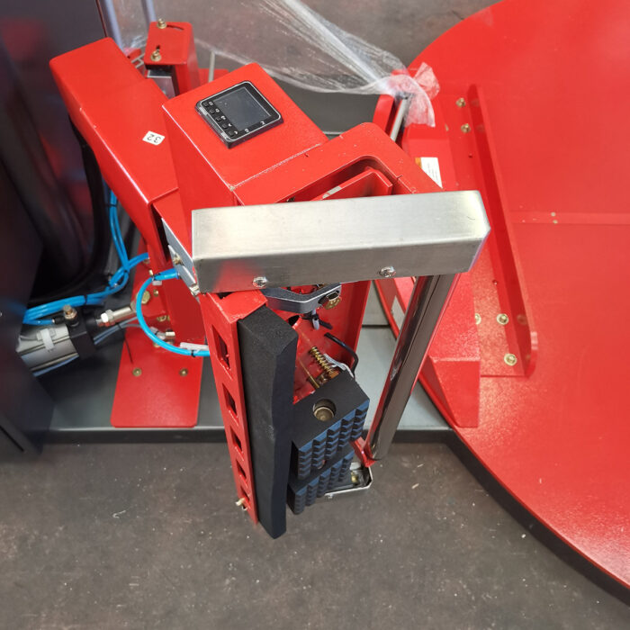 Zbliżenie na Automatyczną owijarkę palet Ultra Wrap 2000 BF, charakteryzującą się czerwoną, przemysłową konstrukcją z metalowymi częściami, widocznym okablowaniem i cyfrowym wyświetlaczem, ustawioną na pasującej czerwonej podstawie na szarej podłodze.