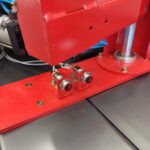 Zbliżenie na dwa czerwone i srebrne czujniki z przewodami i śrubami, zamontowane na czerwonej metalowej podstawie maszyny Automatyczna owijarka palet Ultra Wrap 2000 BWF, umieszczona na szarej i czerwonej platformie przemysłowej.