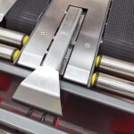 Zbliżenie na przenośnik ze stali nierdzewnej z czarnymi pasami, żółtymi rolkami i metalowym rozdzielaczem - idealny do integracji z Półautomatyczną zaklejarką Random Tape 50 TBB w celu usprawnienia procesów pakowania.