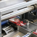 Zbliżenie na Automatyczną pakowarkę flow-pack SR750 zgrzewającą plastikowy pojemnik z truskawkami przezroczystą folią w warunkach przemysłowych.