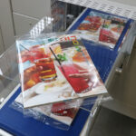 Magazyn przedstawiający ręce trzymające kolorowe napoje jest owijany w przezroczysty plastik przez Automatyczną pakowarkę flow-pack SR550 i umieszczany na niebieskim przenośniku taśmowym, gotowy do pakowania lub dystrybucji.