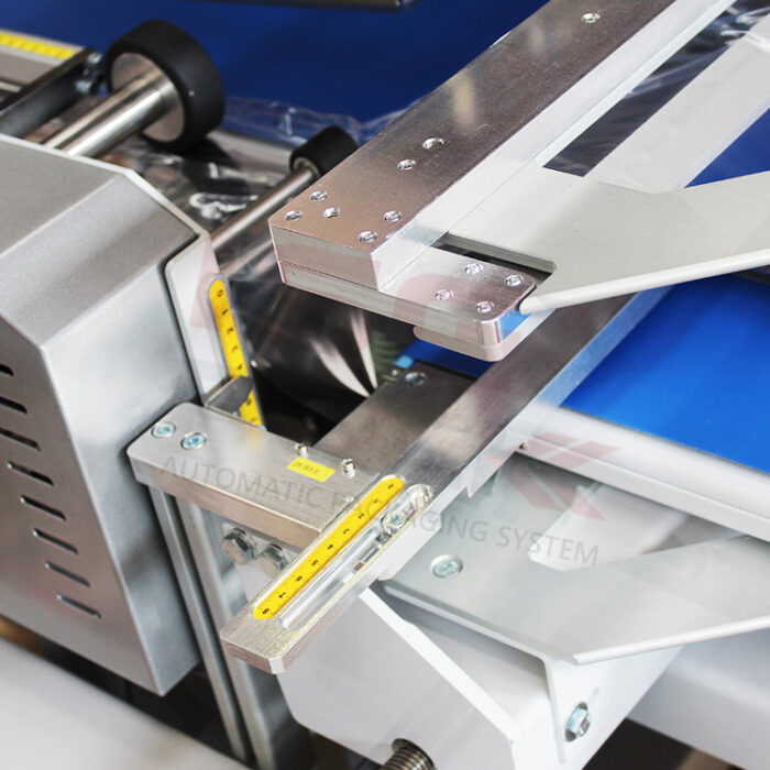 Zbliżenie automatycznej zgrzewarki kątowej Matic Sealer 85 SC z metalowymi ramionami, linijkami pomiarowymi i przenośnikiem taśmowym. Czyste, precyzyjne części pokazują przemysłowy design opakowania.