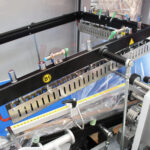 Zbliżenie na przemysłowe maszyny pakujące, przedstawiające Automatyczną zgrzewarkę kątową do folii Matic Sealer 65 E z metalowymi prętami, rolkami, drutami i folią z tworzywa sztucznego przetwarzaną w środowisku fabrycznym.