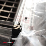 Zbliżenie Zgrzewarki kloszowej do folii Easy Sealer 85, ukazujące jej metalowe części i przezroczystą folię z tworzywa sztucznego z widoczną krawędzią zgrzewającą. Na pierwszym planie widoczne jest logo ISG Pack.