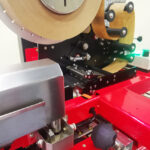 Zbliżenie czerwonej półautomatycznej zaklejarki Easy Paper 50 SB z brązowymi rolkami papieru, wałkami, przekładniami i pokrętłem sterującym, przeznaczonej do półautomatycznego etykietowania produktów.