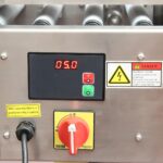 Zbliżenie panelu sterowania Półautomatycznej zaklejarki Eco Tape 50 SB SS: wyświetlacz cyfrowy 05.0, czerwone/zielone przyciski, żółta etykieta ostrzegawcza, czerwony wyłącznik awaryjny i znak ostrzegawczy o niebezpiecznym napięciu.