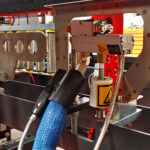 Zbliżenie maszyny przemysłowej z automatyczną formierką Speed Former 50 SB TN HM z niebieskim elastycznym wężem przymocowanym do metalowej złączki, widocznymi etykietami ostrzegawczymi oraz otaczającymi metalowymi częściami i kablami.