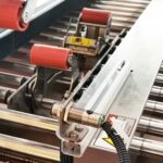 Zbliżenie przemysłowego systemu przenośników z czerwonymi i srebrnymi rolkami, metalowym czujnikiem i okablowaniem zamontowanym obok Automatycznej zaklejarki Auto Tape 50 CF SS do wydajnych zautomatyzowanych procesów produkcyjnych.