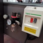 Zbliżenie panelu sterowania Półautomatycznej zaklejarki Random Tape 50 TBB z przełącznikiem Schneider Electric, manometrem i filtrem. Żółta etykieta ostrzega: Upewnij się, że maszyna jest dobrze uziemiona!.