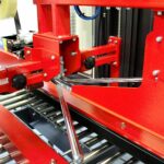 Zbliżenie na Automatyczną zaklejarkę Auto Tape 50 HM, czerwoną automatyczną maszynę pakującą z metalowymi ramionami i rolkami, przeznaczoną do zaklejania produktów na przenośniku.