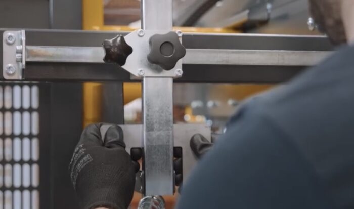 W czarnych rękawiczkach ktoś reguluje metalowe pręty z dużymi pokrętłami na Automatycznej formiarce kartonów SIAT BOXR, skupiając się na swoich dłoniach i komponentach maszyny.