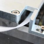 Zbliżenie białego płaskiego kabla połączonego z metalowym elementem Półautomatycznej bandownicy stołowej Eco Strap 501, ukazujące widoczne śruby i precyzyjne wykonanie.