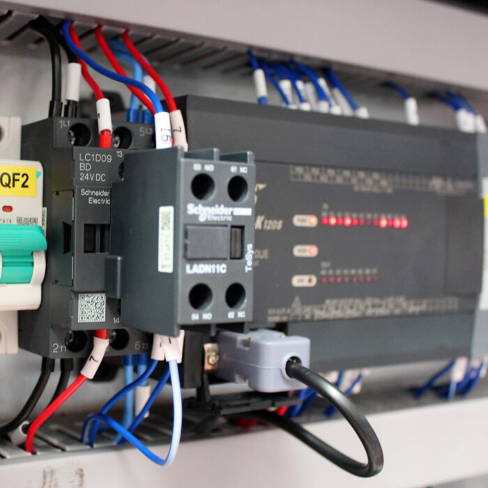 Zbliżenie elektrycznego panelu sterowania Owijarki ramieniowej palet Rotary Wrap 2300 B z przekaźnikiem Schneider Electric, oznakowanym okablowaniem i zaciskami oraz czerwonymi diodami LED wewnątrz szarej obudowy.