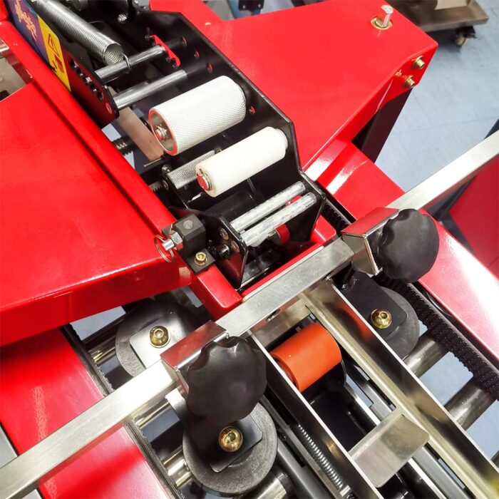 Zbliżenie na Półautomatyczną zaklejarkę Small Tape 50 SB, czerwoną maszynę przemysłową z rolkami, pokrętłami i metalowymi częściami używanymi do przetwarzania lub przenoszenia materiałów w produkcji.