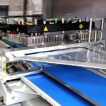 Zbliżenie na Automatyczną zgrzewarkę kątową do folii Matic Sealer 65 E, pokazujące jej niebieski przenośnik taśmowy, przezroczystą folię z tworzywa sztucznego i metalowe części przeznaczone do zgrzewania i pakowania produktów.