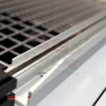 Zbliżenie metalowej siatki i systemu szyn ze Zgrzewarki kloszowej do folii Eco Sealer 45, podkreślające przecinające się metalowe elementy. Logo ISGPACK pojawia się w lewym dolnym rogu.