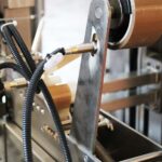 Zbliżenie metalowego ramienia, brązowej rolki taśmy i czujnika z czarnymi kablami na Automatycznej zaklejarce Auto Tape 50 CF SS - przemysłowej maszynie pakującej.