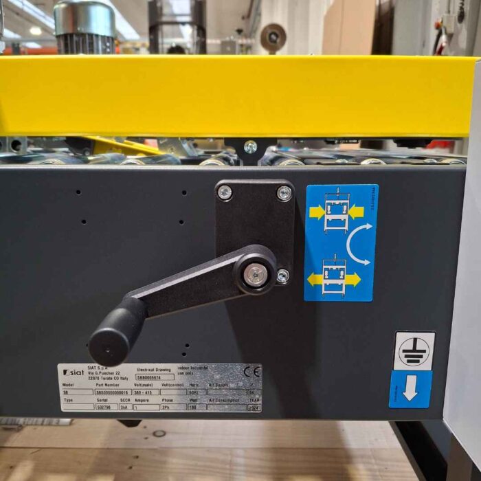 Zbliżenie Półautomatyczna zaklejarka kartonów SIAT S8 posiada czarną korbę ręczną, żółtą barierkę ochronną, niebieską naklejkę instruktażową ze wskazówkami dotyczącymi korby oraz metalową tabliczkę z informacjami technicznymi poniżej korby.