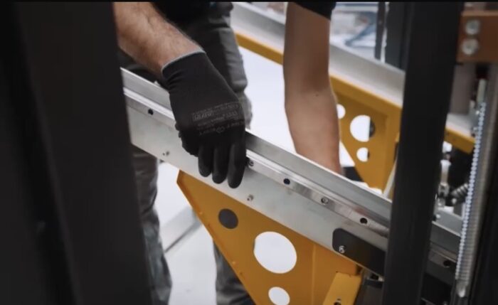 Dwie osoby w czarnych rękawicach montują lub regulują metalową i żółtą część Automatycznej formiarki kartonów SIAT BOXR, co wskazuje na środowisko techniczne lub produkcyjne.