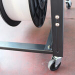 Zbliżenie na metalowy wózek Półautomatyczna bandownica stołowa Eco Strap 501 z kółkami, trzymający dużą białą szpulę taśmy. Wózek stoi na czerwonawej podłodze, ukazując część szpuli i kółka.