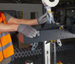 Osoba w szarych rękawicach i pomarańczowej kamizelce ochronnej reguluje czarną część maszyny na półautomatycznej zaklejarce kartonów NASTRO-SB - R5-R6. Powyżej znajduje się rolka taśmy samoprzylepnej SIS, a w tle ułożone są kartony.