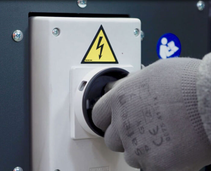 Dłoń w rękawiczce przekręca duży czarny przełącznik elektryczny na białym panelu z żółtym znakiem ostrzegawczym wysokiego napięcia, przygotowując półautomatyczną zaklejarkę kartonów NASTRO-SB L5-L6-L8 do pracy.