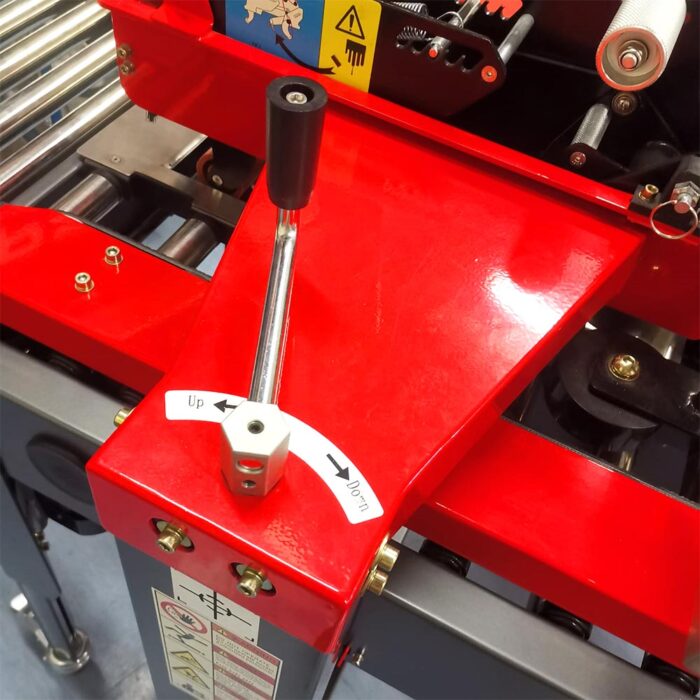 Zbliżenie na półautomatyczną zaklejarkę Small Tape 50 SB, czerwoną maszynę przemysłową wyposażoną w metalową dźwignię z napisem W górę i W dół, z rolkami i ostrzegawczymi etykietami bezpieczeństwa widocznymi w tle.