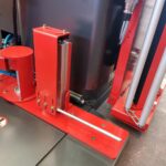 Zbliżenie automatycznej owijarki palet Ultra Wrap 2000 BWF w kolorze czerwonym, pokazujące metalowe pręty, śruby i pionowy mechanizm szynowy na metalowej powierzchni wśród różnych komponentów i rur.