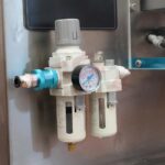 Zbliżenie regulatora powietrza Pionowa maszyna pakująca Mini Vert 240 PP z manometrem, filtrem i pochłaniaczem wilgoci na metalowym panelu. Manometr pokazuje odczyty ciśnienia za pomocą dołączonych przewodów.