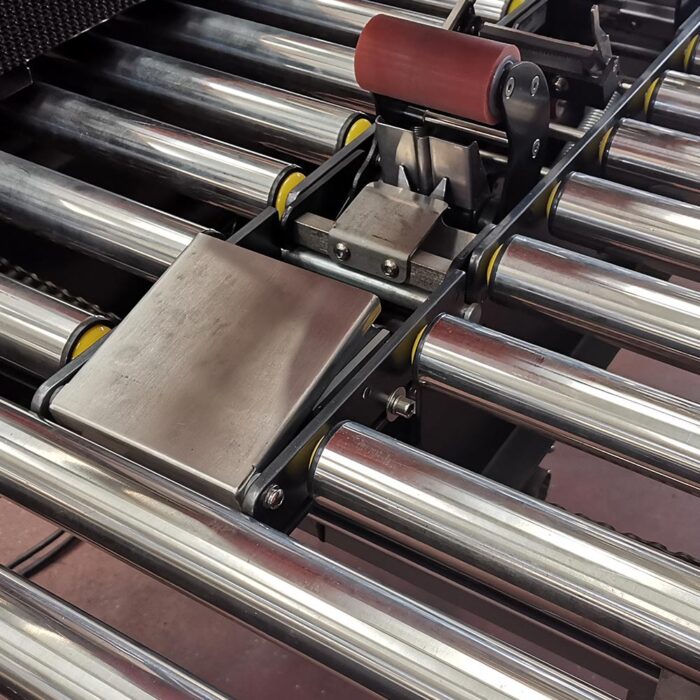 Zbliżenie systemu przenośników z błyszczącymi metalowymi rolkami, z mechanizmem Półautomatyczna zaklejarka Random Tape 50 SB powyżej - obok czerwonej gumowej rolki i metalowych elementów przetwarzających przedmioty na linii.