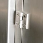 Zbliżenie metalowego zawiasu, takiego jak w Automatycznej zaklejarce Auto Tape 50 CF SS, przymocowanego do dwóch paneli ze stali nierdzewnej, pokazującego mechanizm i cztery śruby mocujące.