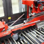 Zbliżenie na Automatyczną zaklejarkę Matic Tape 50 CF HM, zautomatyzowaną maszynę pakującą z czerwonymi metalowymi częściami, rolkami i zmechanizowanymi ramionami do przenoszenia materiałów w środowiskach przemysłowych.