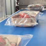 Kilka pudełek pomidorów owiniętych w przezroczysty plastik znajduje się na niebieskim przenośniku taśmowym, gotowych do zamknięcia przez Automatyczną pakowarkę flow-pack SR750 w nowoczesnym zakładzie pakowania żywności.