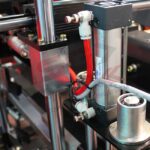 Zbliżenie elementów maszyny przemysłowej - metalowych prętów, siłownika pneumatycznego, węży powietrznych i okablowania - występujących w zautomatyzowanym systemie napełniania lub montażu Automatyczna zaklejarka Matic Tape 50 CF.
