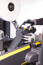 W rękawicach odpornych na przecięcia osoba instaluje lub reguluje rolkę folii opakowaniowej na SIAT SR20 - półautomatycznej zaklejarce kartonów, ostrożnie prowadząc folię przez rolki maszyny.