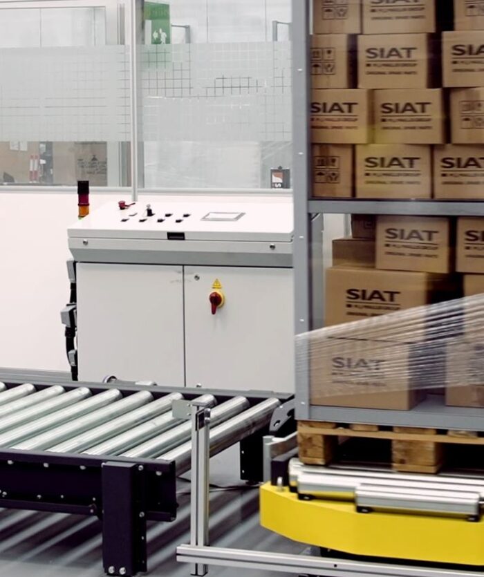 System przenośników, panel sterowania i paleta skrzynek SIAT owiniętych folią podkreślają wydajność Automatycznej owijarki palet ProWrap F/A w środowisku magazynu przemysłowego.