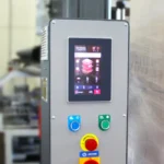 Zbliżenie przemysłowego panelu sterowania dla Ekonomicznej owijarki do palet E-wrap 1000A, z cyfrowym ekranem dotykowym z elementami sterującymi systemu, dwoma zielonymi przyciskami i czerwonym przyciskiem AWARYJNYM; sprzęt wydaje się zamazany w tle.