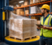 Pracownik w żółtym kasku i kamizelce ochronnej używa tabletu do nadzorowania Automatycznej Owijarki do Palet skutecznie owijającej folię wokół pudełek na palecie w magazynie. W tle widoczne są półki z kolejnymi pudełkami.